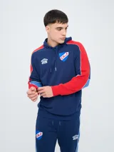 Canguro deportivo Umbro del Club Nacional de Football, modelo Nexo para hombre. Diseño en color azul marino con mangas rojas y detalles en celeste y blanco, capucha con cordón ajustable y bolsillo tipo canguro.