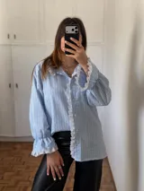 Camisa de rayas verticales celestes y blancas, con cuello alto y cierre frontal de botones. Presenta detalles de puntilla blanca en el cuello, a lo largo de la botonera y en los puños. Tiene mangas largas con terminación acampanada y diseño holgado.