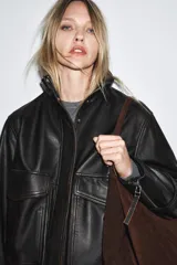 Chaqueta bomber de cuero negro con cuello alto, manga larga con cremalleras metálicas, bolsillos de plastrón con solapa y botón en delantero, bajo acabado en elástico y cierre frontal con cremallera y botones a presión ocultos por solapa.