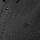 Camisa de manga larga color gris oscuro con textura tipo espiga. Tiene cuello clásico, cierre frontal con botones y un pequeño logo bordado en el pecho. Corte recto.