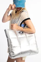 Bolso shopper plateado acolchado, con diseño puffer y costuras a cuadros. Cuenta con tiras para el hombro y un parche ovalado negro y blanco con las iniciales dl en el centro.