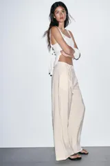 Pantalón de tiro alto en color beige, con diseño de pierna ancha y pinzas frontales. Cuenta con bolsillos laterales, cierre frontal con cremallera y gancho metálico, y un lazo ajustable en la cintura del mismo tejido.