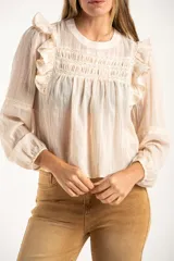 Blusa de color arena con diseño romántico, presenta volados en los hombros, detalles de encaje en el pecho y mangas largas con puños elásticos.