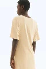 Vestido corto de punto color beige, con cuello redondo y mangas cortas. Presenta un nudo fruncido en el lateral que crea un efecto drapeado y abertura en el ruedo.