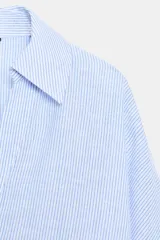 Camisa de mujer color celeste con finas rayas verticales blancas. Presenta un corte amplio y holgado, cuello solapa con escote en pico y mangas anchas que terminan en puño. Cierre frontal con botones.