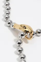 Collar de estilo lariat con cadena de bolas plateada y detalles dorados. Presenta un dije en forma de cabeza de serpiente que funciona como pasador y un terminal en punta que simula la cola. Está confeccionado principalmente en latón con acabado metálico bicolor.