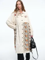 Chaqueta larga de diseño patchwork en tonos grises, con combinación de texturas que incluyen paneles de tela lisa, tela texturizada blanca y paneles con estampado floral en relieve. Presenta cierre frontal con botones, cuello camisero y bordes deshilachados.