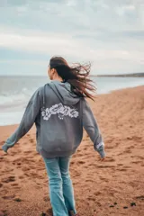 Hoodie gris con capucha, cordones ajustables, mangas largas y puños elásticos. Presenta un estampado de flores blancas en la espalda y un bordado en la manga.