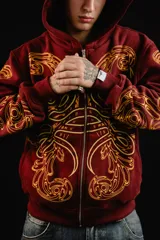 Hoodie con cierre frontal, color bordó, con estampado serigráfico en color dorado que recorre el frente y las mangas con un diseño inspirado en hojas de acanto. Cuenta con bolsillos laterales y cierre metálico.