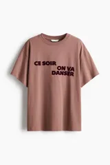 Remera oversized de punto de algodón, de color marrón, con cuello redondo ribeteado, hombros ligeramente caídos, mangas cortas y estampa frontal con texto en contraste.