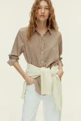 Camisa de manga larga con estampado de rayas verticales en tonos marrón y blanco. Presenta cuello clásico, cierre frontal con botones, puños abotonados y bajo redondeado.