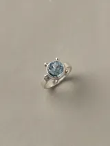Anillo de plata 925 con piedra central de topacio azul facetado en talla brillante de 8 mm, rodeada de pequeñas esferas decorativas.