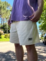Bermuda color crema, de algodón, con calce relajado que cae por encima de la rodilla y cintura elastizada. Presenta un parche rectangular negro con el texto "DROPZ" en blanco en la pierna izquierda.