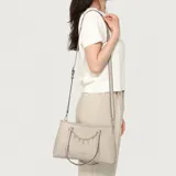 Cartera Guess modelo Angelica de color gris, con diseño estructurado de símil cuero, doble asa superior y correa ajustable. Presenta detalles de herrajes plateados, un charm colgante con las letras de la marca y el logo frontal impreso.