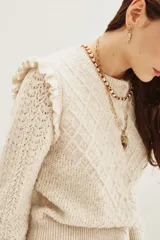 Cardigan de punto color beige con textura calada, cuello redondo y detalle de volados en los hombros. Presenta cierre frontal y puños acanalados.