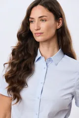 Camisa celeste rayada de manga corta, con cuello camisero y cierre frontal con botones. Presenta un corte entallado y un pequeño detalle de placa metálica en el borde inferior.