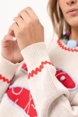 Sweater tejido color crudo con cuello redondo y mangas largas. Presenta dos peces rojos con detalles en azul bordados en el frente. Tiene ribete ondulado rojo en cuello, puños y ruedo.