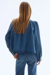 Campera de sarga de mezclilla azul, con cuello elevado, cierre frontal de botones y canesú fruncido. Tiene mangas largas abullonadas con puños ajustados y bolsillos laterales insertados.