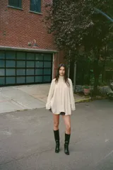 Top tipo capa de color beige, con cuello subido y cierre posterior con botones ocultos. Presenta un diseño fluido con bajo asimétrico y mangas acampanadas.