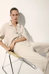 Pantalón sastrero color beige de tiro alto, confeccionado en mezcla de lino y lyocell. Presenta diseño de perneras rectas con pliegues frontales y traseros, bolsillos al bies y trabillas para cinturón.