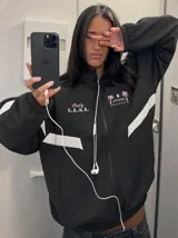 Campera deportiva negra con cuello alto, cierre frontal completo y franjas blancas en las mangas. Presenta estampados en color rosado en el pecho, incluyendo la inscripción "Only F.A.N.S." y gráficos de palmeras con estrellas.