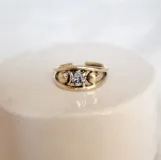 Anillo de bronce dorado con diseño abierto y una zirconia blanca engarzada en el centro. Dos pequeñas esferas de bronce decoran cada lado de la piedra.