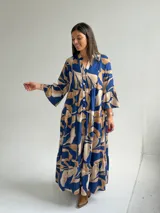 Vestido largo azul con estampado de hojas en tonos beige y blanco, escote en V con botones frontales, mangas largas amplias y diseño escalonado.