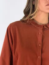 Camisa de pana color chocolate, con cuello alto tipo mao con volados, cierre frontal de botones y mangas largas con puños abotonados.