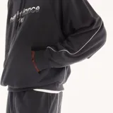 Canguro New Balance Trackside de color negro, confeccionado en felpa francesa de algodón y poliéster reciclado. Presenta mangas raglán con ribetes blancos en contraste, capucha ajustable con cordón, bolsillo tipo canguro delantero y puños y dobladillo acanalados. Incorpora el logotipo y emblema gráfico de la marca bordado y estampado en el pecho.