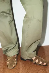 Sandalia plana tipo cangrejera en serraje color marrón claro, con tiras entrelazadas en la parte delantera y cierre de hebilla en el tobillo. Tiene punta redonda y un tacón bajo de 2 cm.