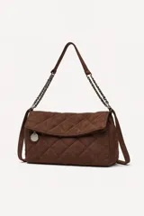 Cartera tipo bandolera de gamuza marrón con diseño acolchado de rombos. Cuenta con una solapa frontal, un dije metálico circular colgante y una correa combinada de cadena metálica y gamuza.