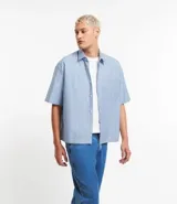 Camisa masculina oversized boxy de manga corta, confeccionada en tejido oxford a rayas verticales celestes y blancas. Cuenta con cuello inglés, abotonadura frontal y bolsillo de parche en el pecho.