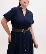 Vestido midi chemise azul marino de manga corta, con cuello padre, botonadura frontal completa con botones metalizados y pliegues en la falda. Incluye un cinturón marrón.