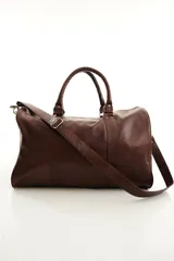 Bolso de viaje color marrón, confeccionado en cuero vacuno, con doble asa de mano y correa ajustable y desmontable. Cuenta con herrajes metálicos y costuras reforzadas.