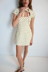 Vestido corto de color blanco con estampado de lunares negros. Posee cuello redondo con lazada, escote cuadrado en la parte delantera y abertura tipo gota con lazo en la espalda. Mangas cortas y silueta entallada.