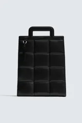 Bolso tipo shopper de diseño acolchado con patrón de cuadros, confeccionado en material sintético negro. Presenta asas de mano integradas en la estructura superior y un detalle de borla colgante con herraje metálico.
