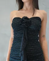 Vestido corto strapless negro con diseño drapeado en toda la prenda, detalle de aplique floral en el centro del escote y caída en cascada con volados frontales. Presenta pequeños puntos brillantes distribuidos por toda la superficie.