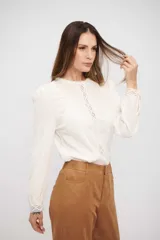 Blusa de color crudo con detalles de encaje vertical en el frente y en los puños. Presenta mangas largas con volumen y cuello redondo.