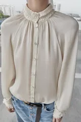 Blusa fluida de color blanco roto o crema, con cuello alto fruncido y manga larga con puños elásticos tipo nido de abeja. Presenta un frente abotonado con botones pequeños y brillantes.