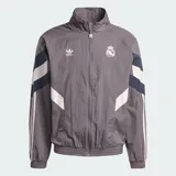 Chaqueta deportiva Adidas del Real Madrid en color gris, con diseño de bloques en blanco y azul marino, cuello alto, cierre frontal y las icónicas tres tiras en las mangas.