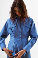 Camisa azul de algodón con finas rayas blancas verticales, cuello mao, manga larga y detalle de pinzas y botones en la zona de la cintura. Posee dos bolsillos de parche con tapa en el pecho.