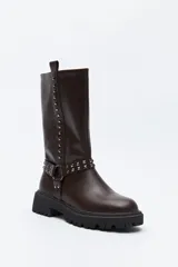 Bota de caña media estilo biker, confeccionada en cuero sintético color marrón oscuro. Presenta una suela track robusta, detalle de tachas metálicas verticales a lo largo de la caña y un arnés decorativo con argolla metálica en el tobillo.