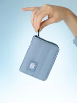 Monedero rectangular de lona color azul grisáceo, con cierre perimetral y costuras decorativas verticales. Incluye un parche frontal con el logo de la marca.