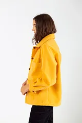 Camisa color mostaza de paño, corte oversize, con cuello clásico, cierre frontal con botones, mangas largas con puños abotonados y dos bolsillos de parche en el pecho.