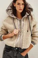Campera de estilo híbrido con cuerpo de corderito sintético color beige y panel frontal central con cierre y capucha de tela técnica metalizada en tono grisáceo. Presenta cordones ajustables en la capucha con diseño estampado y puños con detalle de rayas.