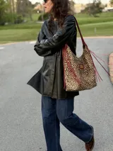 Cartera tipo hobo de cuero con estampado animal print de leopardo, con tiras y detalles en cuero rojo con flecos y parche rectangular con logo grabado en la parte frontal.
