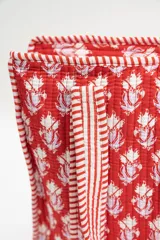Bolso tipo tote acolchado con estampado floral en tonos rojizos, rosados y verdes. Presenta asas de mano con diseño de rayas verticales en color rojo y blanco, y ribetes a juego en los bordes.
