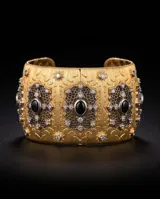 Brazalete rígido de bronce con baño dorado de la línea Imperial, diseño texturizado con apliques metálicos ornamentales en tono plateado, incrustaciones de circonias y piedras negras ovales centrales.