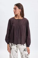 Blusa de color marrón con diseño de alforzas en la parte superior, cuello redondo y cierre frontal con botones. Presenta mangas largas con puños elásticos y un corte holgado con frunces bajo el canesú.