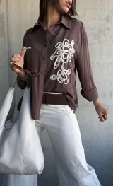 Camisa de manga larga color chocolate, con cuello clásico y cierre frontal de botones. Presenta un bordado decorativo en color crudo en el frente con diseño de formas orgánicas.